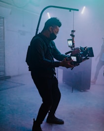 A person wearing a face mask operates a professional camera with a stabilizer in a dimly lit room filled with fog. The room has industrial elements, including metallic surfaces and fluorescent lights overhead, giving it a cinematic atmosphere.