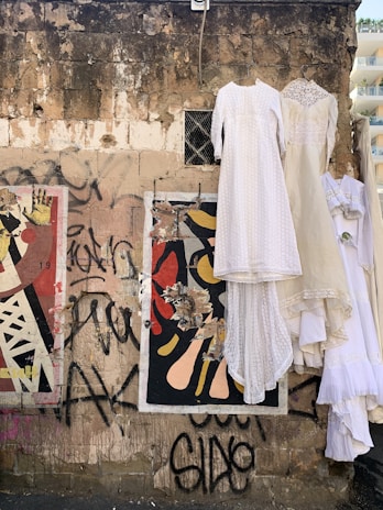 A worn and textured brick wall is adorned with colorful graffiti and torn posters. Elegant, white vintage dresses hang from metal hangers attached to the wall, creating a stark contrast. The scene reflects an intersection of urban decay and retro fashion.