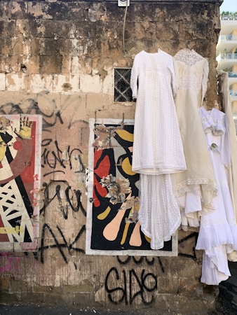 A worn and textured brick wall is adorned with colorful graffiti and torn posters. Elegant, white vintage dresses hang from metal hangers attached to the wall, creating a stark contrast. The scene reflects an intersection of urban decay and retro fashion.