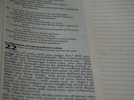 A close-up view of an open page from a book, likely a Bible, featuring printed text with headings and numbered verses. The right-hand side shows a blank lined section.