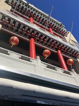 A building facade with traditional architectural elements featuring three red pillars and several red and gold Chinese lanterns hanging from the upper levels. The structure includes horizontal beams and intricate designs, all set against a clear blue sky.