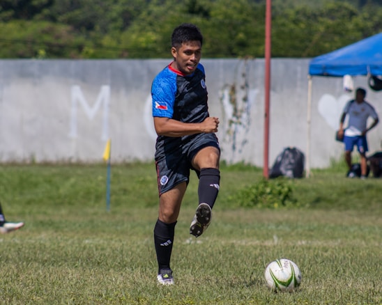 A soccer player wearing a blue and black kit is actively engaged in a game, preparing to kick a green and white soccer ball on a grass field. In the background, a fence and a blue tent are visible along with another person standing casually.