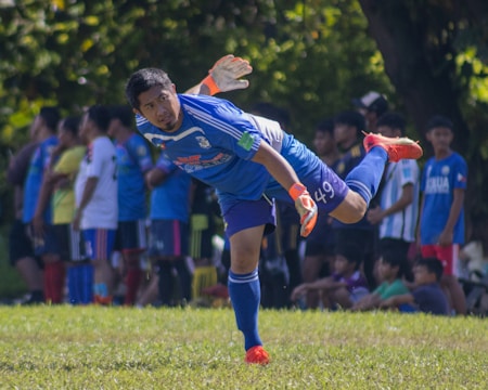 A soccer player in a blue jersey and shorts is actively engaged in the game, positioned on one leg as he leans forward with his other leg extended behind him. He wears a pair of gloves and bright red shoes. In the background, a group of spectators and other players are standing and watching, with trees and greenery providing a natural setting.
