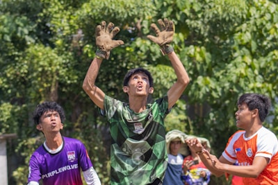 A group of young men appears to be playing a game of soccer outdoors. The central figure is wearing a green and black checkered shirt, raising his arms as if about to catch or block a ball. He is wearing gloves and seems focused on the action above. On either side of him, other participants watching intently, wearing purple and orange shirts respectively. The background features lush green trees, indicating an outdoor setting.