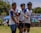 Three young men are standing outdoors, wearing sports jerseys and athletic shorts. They appear to be on a playing field, participating in a sporting event. The background includes a banner, a tent, and some spectators. There are trees and clear skies also visible in the background.
