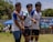 Three young men are standing outdoors, wearing sports jerseys and athletic shorts. They appear to be on a playing field, participating in a sporting event. The background includes a banner, a tent, and some spectators. There are trees and clear skies also visible in the background.