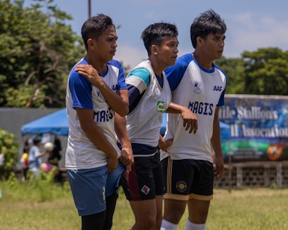 Three young men are standing outdoors, wearing sports jerseys and athletic shorts. They appear to be on a playing field, participating in a sporting event. The background includes a banner, a tent, and some spectators. There are trees and clear skies also visible in the background.