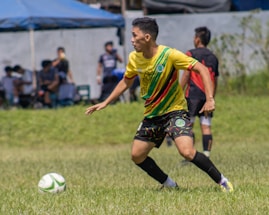 A soccer player wearing a yellow jersey with green and red accents is running on a grassy field. He is focused on controlling a white and green soccer ball. In the background, there are other players and spectators, along with a blue tent.