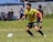 A soccer player wearing a yellow jersey with green and red accents is running on a grassy field. He is focused on controlling a white and green soccer ball. In the background, there are other players and spectators, along with a blue tent.