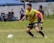 A soccer player wearing a yellow jersey with green and red accents is running on a grassy field. He is focused on controlling a white and green soccer ball. In the background, there are other players and spectators, along with a blue tent.