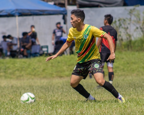 A soccer player wearing a yellow jersey with green and red accents is running on a grassy field. He is focused on controlling a white and green soccer ball. In the background, there are other players and spectators, along with a blue tent.