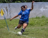 A person in a blue sports jersey is actively kicking a soccer ball with their left foot on a grassy field. Another person is partially visible in the background, also wearing sports attire. A yellow corner flag is positioned to the left, and a concrete wall with a faded arrow marking is in the background.