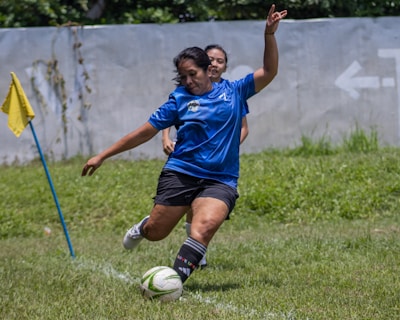 A person in a blue sports jersey is actively kicking a soccer ball with their left foot on a grassy field. Another person is partially visible in the background, also wearing sports attire. A yellow corner flag is positioned to the left, and a concrete wall with a faded arrow marking is in the background.