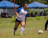 A female soccer player is in motion on a grassy field, preparing to kick a ball. She is wearing a white and blue jersey, black shorts, and bright green cleats. A tent with people sitting underneath is visible in the background.