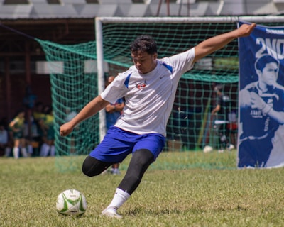 A soccer player in action is about to kick a ball on a grassy field. The player is wearing a white jersey, blue shorts, and black leggings. A soccer goal with a green net is visible in the background, along with spectators and another banner showing an athlete.