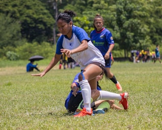 A group of young people engaged in a soccer game on a grassy field, with one player in a white and blue uniform falling while trying to maintain balance. Other players in blue jerseys and spectators are visible in the background, with a lush green environment surrounding them.
