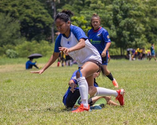 A group of young people engaged in a soccer game on a grassy field, with one player in a white and blue uniform falling while trying to maintain balance. Other players in blue jerseys and spectators are visible in the background, with a lush green environment surrounding them.