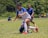 A group of young people engaged in a soccer game on a grassy field, with one player in a white and blue uniform falling while trying to maintain balance. Other players in blue jerseys and spectators are visible in the background, with a lush green environment surrounding them.