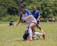 A group of young people engaged in a soccer game on a grassy field, with one player in a white and blue uniform falling while trying to maintain balance. Other players in blue jerseys and spectators are visible in the background, with a lush green environment surrounding them.