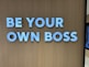 Large white letters spelling 'BE YOUR OWN BOSS' are mounted on a wooden wall, conveying empowerment and independence.