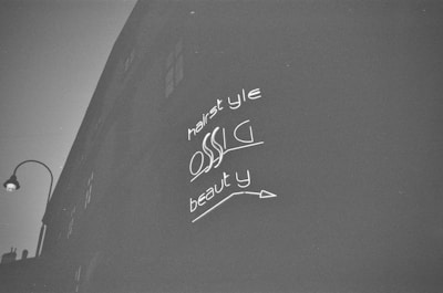 A black and white photograph depicts a dark building facade with bright neon-like signage. The words 'hairstyle', 'OSSC', and 'beauty' are visible, each with distinctive stylization. A vintage street lamp is positioned on the left side.