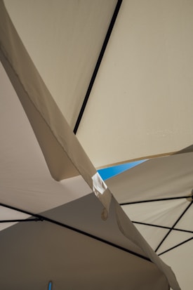 Beige and white fabric panels intersect with black metal poles, creating a geometric pattern. Sunlight gently illuminates the fabric, and a small patch of clear blue sky is visible in the background.