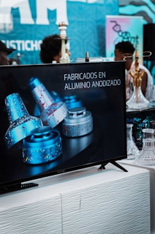 A television screen on a white stand displays an advertisement for products made from anodized aluminum with blue and silver colors featuring a speckled design. In the background, blurred figures of people and decorative items such as hookahs or vases can be seen, suggesting a retail or exhibition setting with a blue and pinkish color theme.