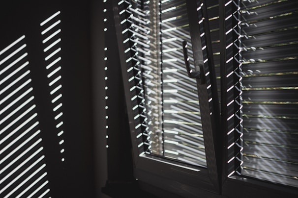 Light filters through horizontal blinds, casting sharp, linear shadows on a wall. The window is partially open, allowing sunlight to highlight the metallic surface of the blinds.