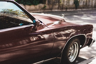 A vintage car with shiny, deep red paint is parked on a street. Reflections of the surroundings are visible on the car's surface, showcasing its polished state. The car's front section, including part of the hood and side mirror, is in focus. The setting is outdoors, with a blurred background of trees and a stone wall.