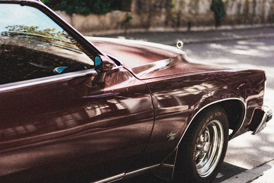 A vintage car with shiny, deep red paint is parked on a street. Reflections of the surroundings are visible on the car's surface, showcasing its polished state. The car's front section, including part of the hood and side mirror, is in focus. The setting is outdoors, with a blurred background of trees and a stone wall.