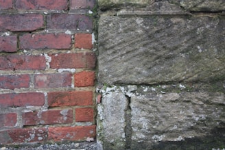 A side-by-side view of two different types of walls, one made of red bricks and the other of large stone blocks. The brick wall appears aged, with visible white mortar and some moss or lichen growth. The stone wall has a rough texture with a mix of earthy tones and is also speckled with lichen.