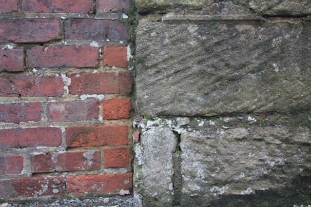 A side-by-side view of two different types of walls, one made of red bricks and the other of large stone blocks. The brick wall appears aged, with visible white mortar and some moss or lichen growth. The stone wall has a rough texture with a mix of earthy tones and is also speckled with lichen.