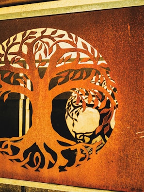 A metal panel features a detailed cutout of a tree with intricate branches and leaves. Behind the panel, a stylized figure reminiscent of a person's head is visible, creating an artistic interplay between the foreground and background.