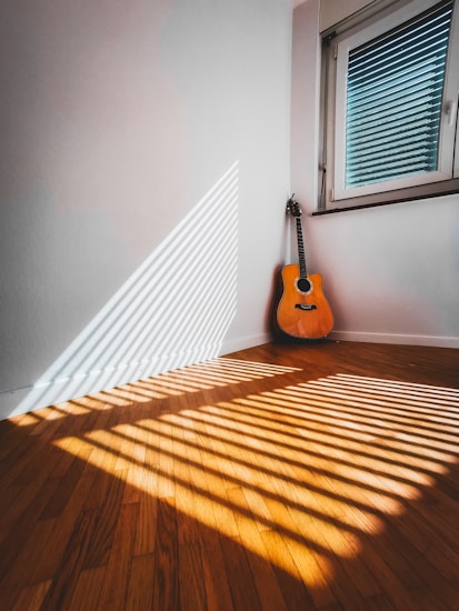 Sunlight streams through window blinds, casting striped shadows on a wooden floor. In the corner of the room stands an acoustic guitar, propped against the wall. The warm tones of the wood and the bright light create an inviting and serene atmosphere.