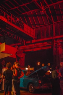A dimly lit industrial space features groups of people gathered around a vintage blue car with its hood open. The area is illuminated by dramatic red lighting, creating a warm atmosphere. The ceiling includes metal beams and rustic structures, adding to the urban and nostalgic setting.