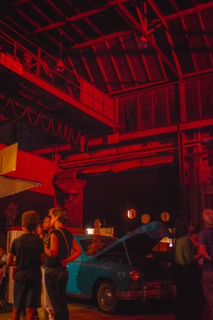 A dimly lit industrial space features groups of people gathered around a vintage blue car with its hood open. The area is illuminated by dramatic red lighting, creating a warm atmosphere. The ceiling includes metal beams and rustic structures, adding to the urban and nostalgic setting.