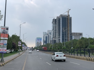 A street view with a wide road lined by tall lamp posts and trees on one side. Modern high-rise buildings under construction are visible in the distance, with a large billboard displaying an advertisement. Several cars, including a white car in the foreground, are driving on the road.