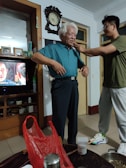 An elderly man with white hair is standing in a room wearing a blue shirt and dark trousers. A younger person is adjusting or helping him with something around his neck. The room contains shelves with various items, including a television showing a scene from a show or movie. A large wall clock is visible above the man. On a table in the foreground, there is a red plastic bag and some household items.