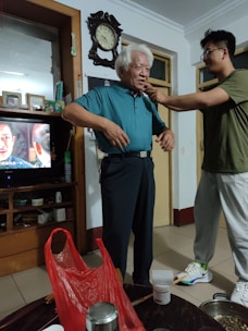 An elderly man with white hair is standing in a room wearing a blue shirt and dark trousers. A younger person is adjusting or helping him with something around his neck. The room contains shelves with various items, including a television showing a scene from a show or movie. A large wall clock is visible above the man. On a table in the foreground, there is a red plastic bag and some household items.