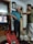 An elderly man with white hair is standing in a room wearing a blue shirt and dark trousers. A younger person is adjusting or helping him with something around his neck. The room contains shelves with various items, including a television showing a scene from a show or movie. A large wall clock is visible above the man. On a table in the foreground, there is a red plastic bag and some household items.