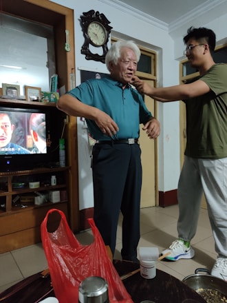 An elderly man with white hair is standing in a room wearing a blue shirt and dark trousers. A younger person is adjusting or helping him with something around his neck. The room contains shelves with various items, including a television showing a scene from a show or movie. A large wall clock is visible above the man. On a table in the foreground, there is a red plastic bag and some household items.