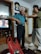 An elderly man with white hair is standing in a room wearing a blue shirt and dark trousers. A younger person is adjusting or helping him with something around his neck. The room contains shelves with various items, including a television showing a scene from a show or movie. A large wall clock is visible above the man. On a table in the foreground, there is a red plastic bag and some household items.