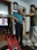 An elderly man with white hair is standing in a room wearing a blue shirt and dark trousers. A younger person is adjusting or helping him with something around his neck. The room contains shelves with various items, including a television showing a scene from a show or movie. A large wall clock is visible above the man. On a table in the foreground, there is a red plastic bag and some household items.