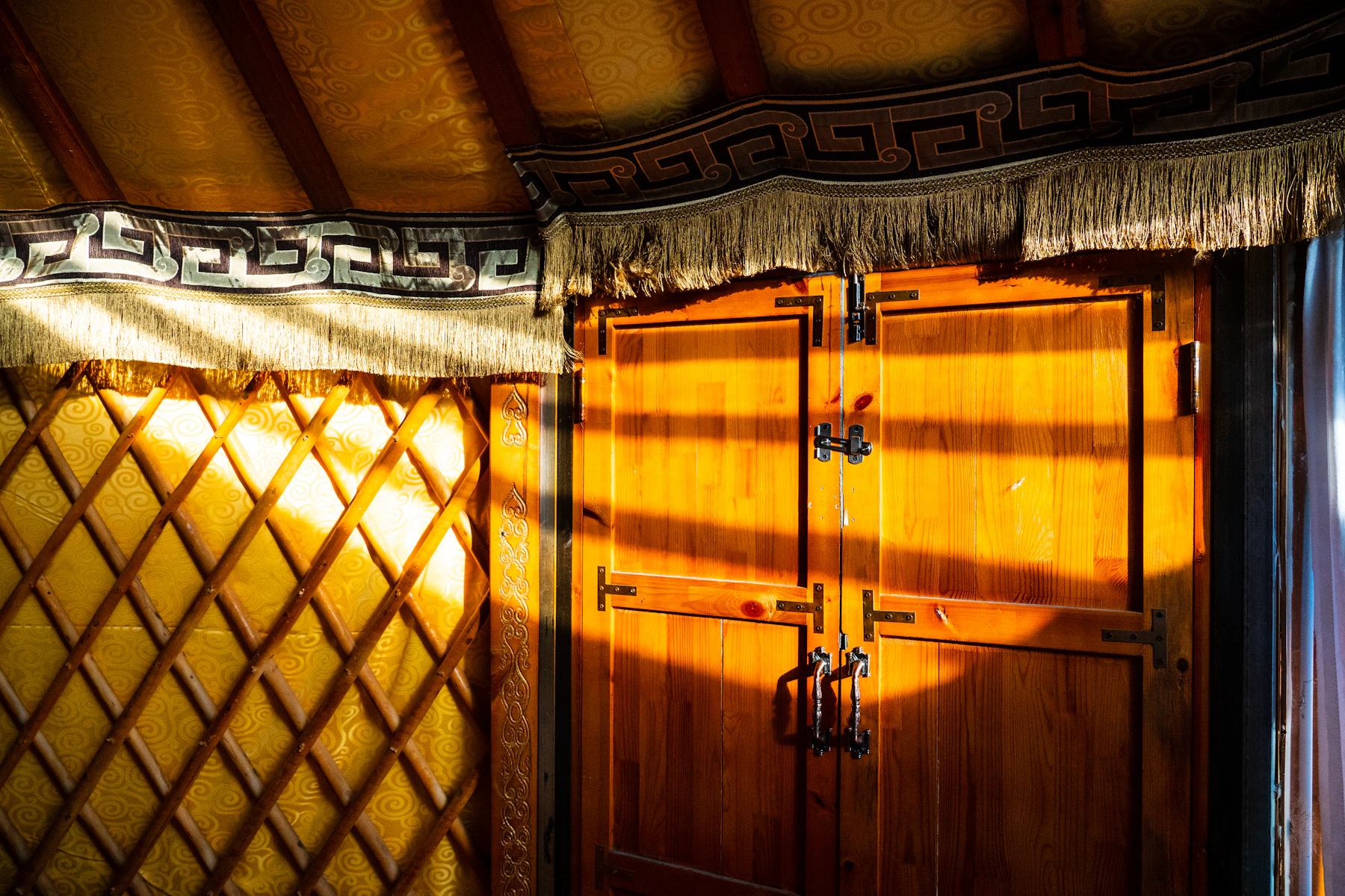 Sunlight on a lattice wall and a painted wooden double door inside a ger, a woven band running above them