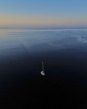 A wide ocean horizon at dawn with a lone sailboat navigating calm waters.