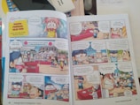 A comic book spread with colorful illustrations and multiple panels featuring animated characters. The scene depicts a cooking contest with chefs and spectators present. Speech bubbles contain dialogue in a foreign language, likely Indonesian. Characters express various emotions, such as excitement, confusion, and surprise, as they discuss aspects of the cooking event. The setting includes tables with dishes and a vibrant outdoor atmosphere.