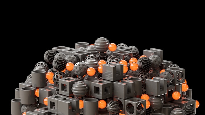 A collection of geometric shapes including cubes, spheres, and cylinders in a variety of perforated and solid designs. The shapes are predominantly in a muted dark gray color, interspersed with bright orange spheres, all placed against a black background.