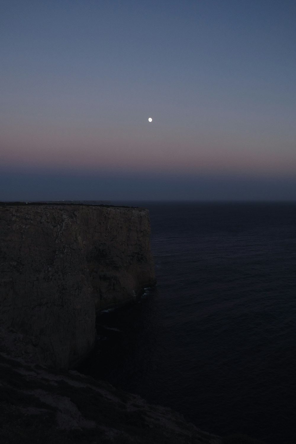 the moon is setting over the ocean on a cliff