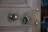 A wooden door featuring an ornate metal keyhole and a polished round door knob. The door has a rich, textured grain, and the hardware reflects light, indicating a metallic composition. The background is blurred, suggesting an interior space with furniture.