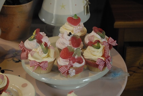 Several decorative cupcakes are placed on a plate, each adorned with various toppings such as gummy candies shaped like hearts and strawberries. The cupcakes appear to have swirled frosting in shades of white and pink, with ribbons tied in bows around the base.